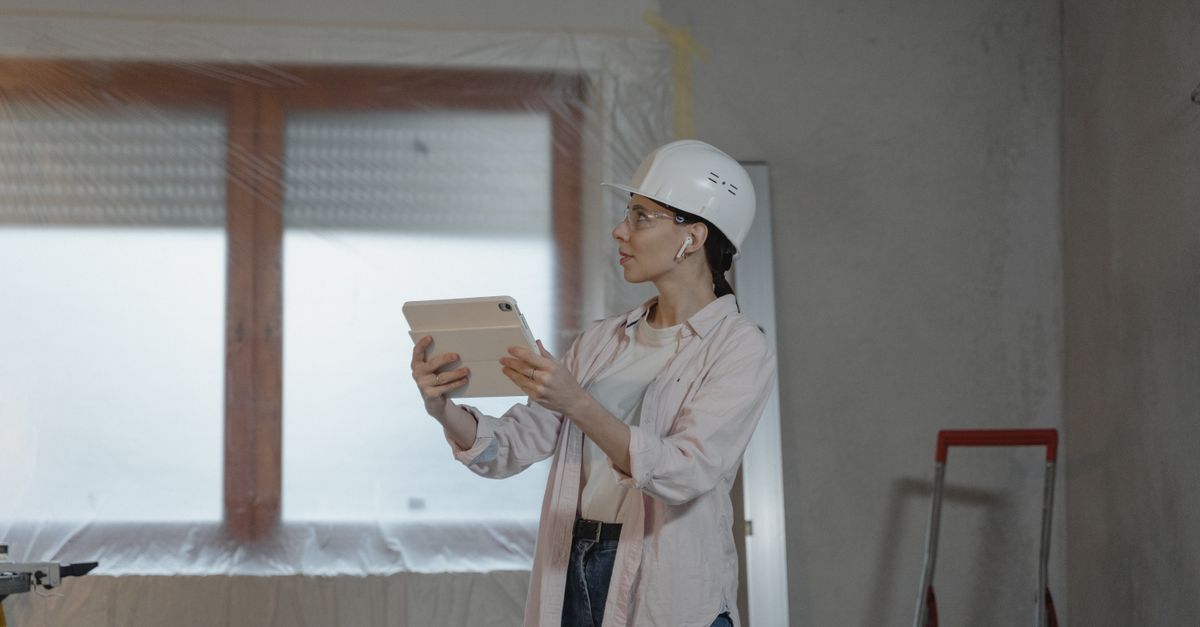 A woman in a hard hat checking a tablet inside an unfinished room