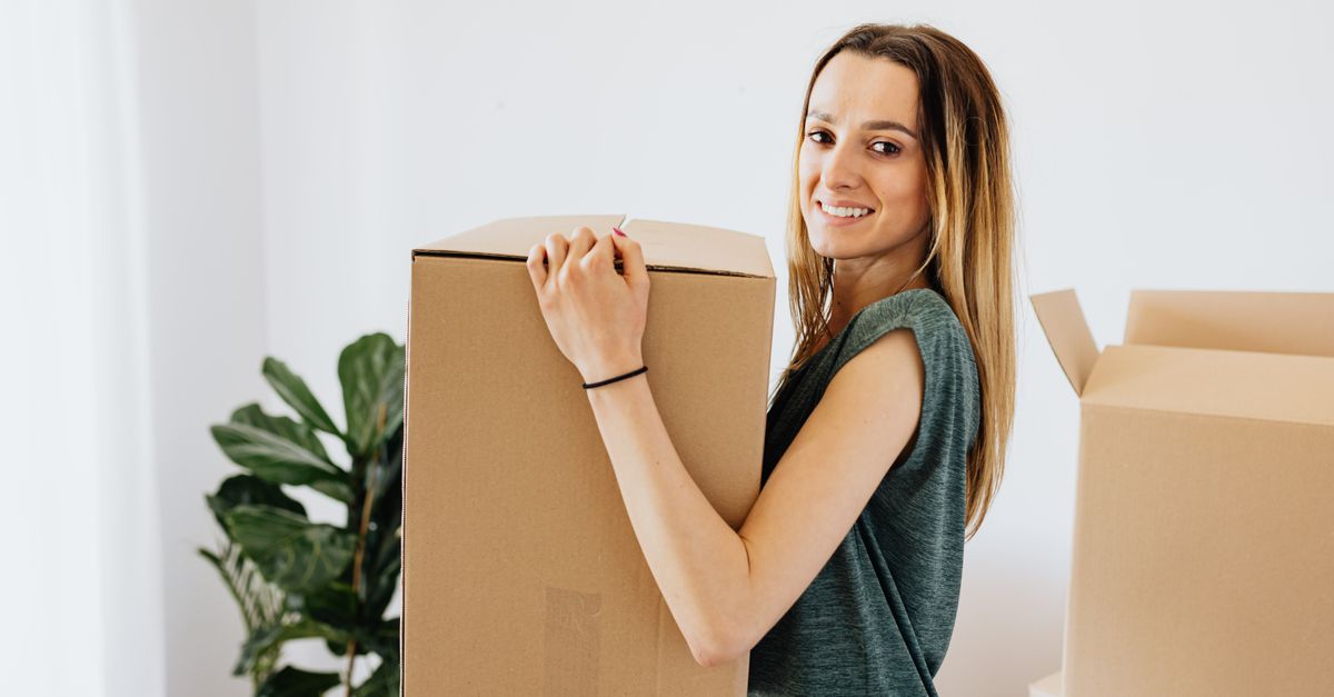 A smiling woman carrying a cardboard moving box in a bright empty apartment